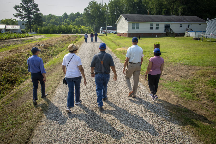 A group of six people walk away from the camera down a gravel path between fields, toward a white house and a parked bus in the distance. The group includes men and women dressed casually, some wearing hats and blue caps. Trees and additional farm structures are visible in the background under a clear sky.