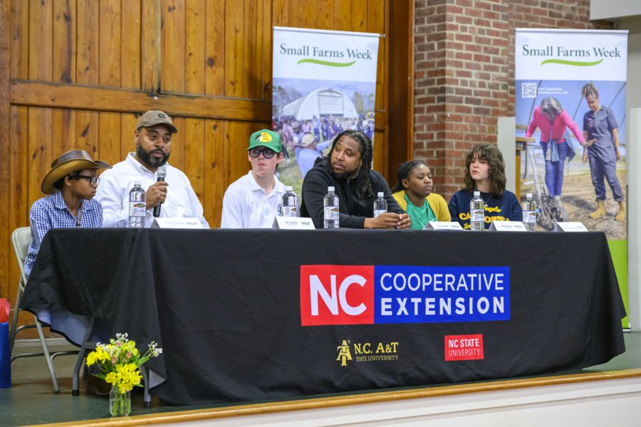 A panel of six diverse speakers, including youth and adults, sits at a table covered with an NC Cooperative Extension banner. Behind them are posters for Small Farms Week, and one man speaks into a microphone while others listen attentively.