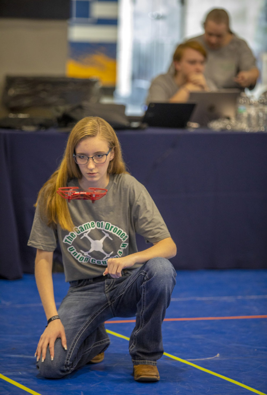 A young person with glasses kneels on a blue floor, focused, wearing a Game of Drones t-shirt, with a small drone hovering near their face. Two people work at a table with laptops in the background.