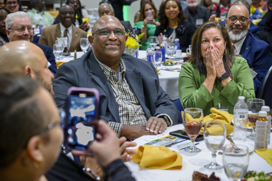 A group of people sits at a round banquet table during a formal event. One man smiles while a woman beside him reacts emotionally with her hands over her mouth. Others around them smile, clap, or take photos, creating a joyful atmosphere.