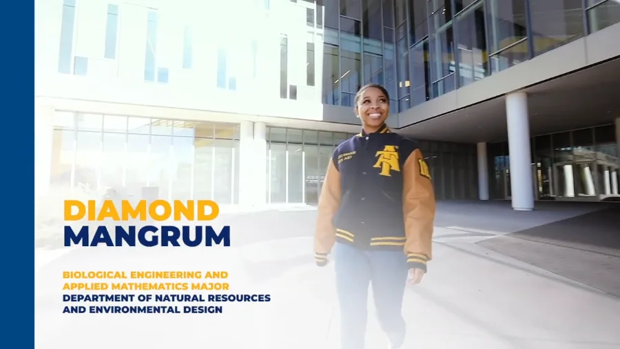 Smiling student wearing a blue and tan varsity jacket with a gold university emblem walks outside a modern glass academic building with tall windows and white pillars.