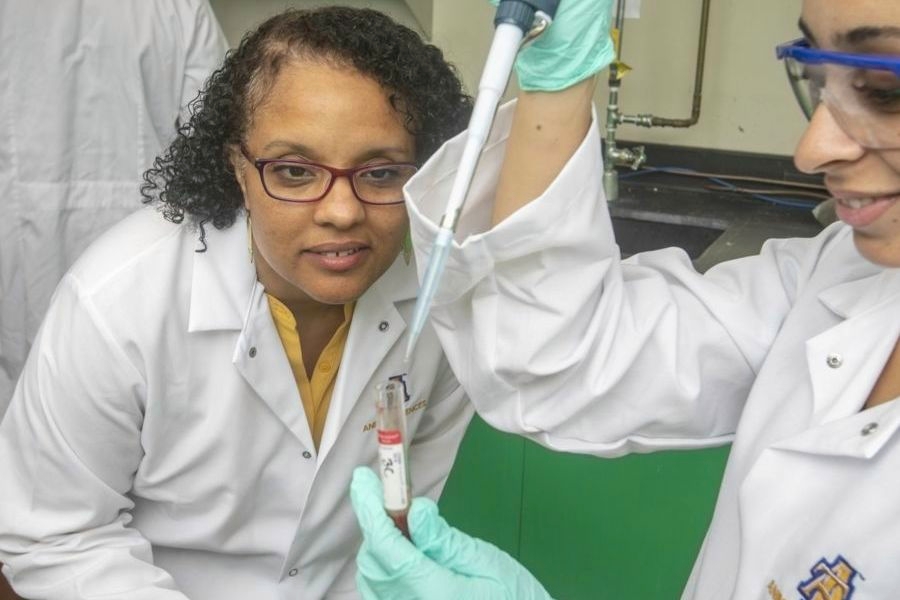 Two scientists in white lab coats working in a laboratory, one using a pipette to transfer liquid into a test tube while the other observes closely.